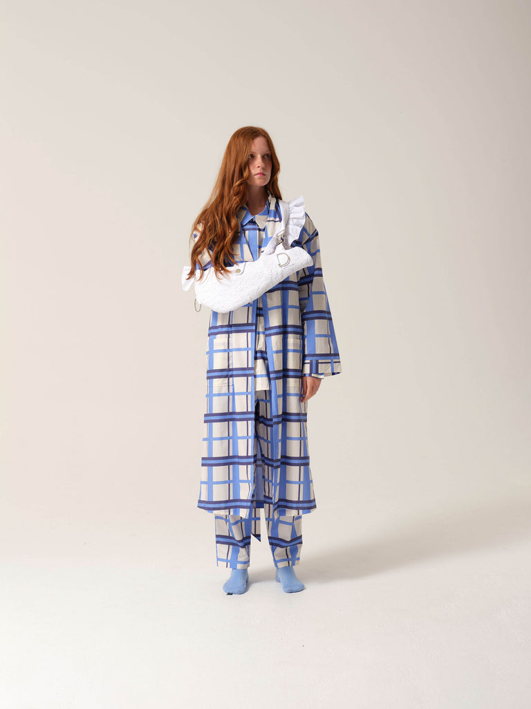 Woman wearing blue and white checkered pyjamas with a white embroidered arm sling against a studio ackground looking away from the camera.