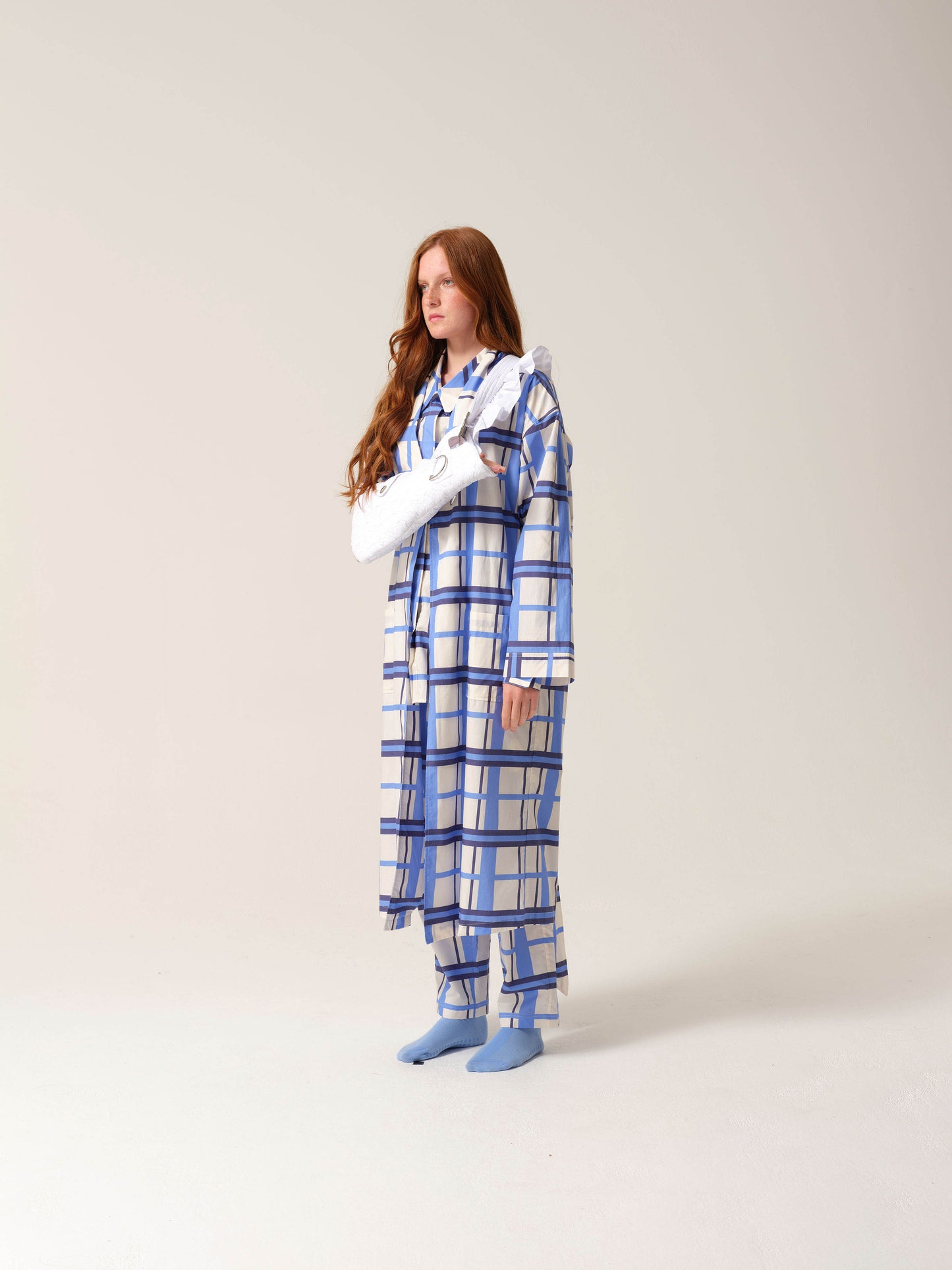 Woman wearing blue and white checkered pyjamas with a white embroidered arm sling against a studio ackground turned to face away from the camera.