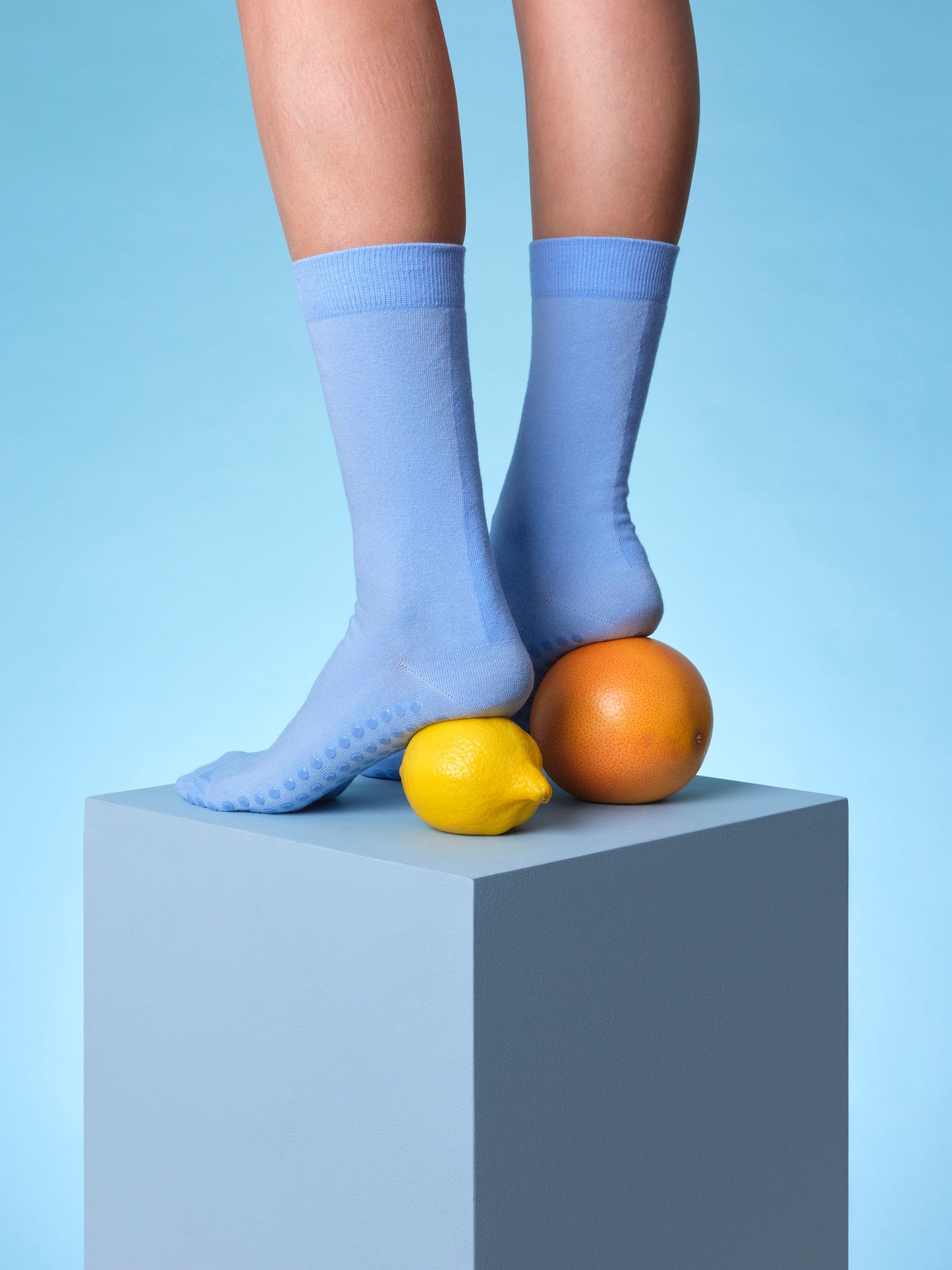 Pair of legs standing on blue plinth in pair of cornflower blue hospital socks with non-slip textured grips soles photographed with lemon and orange under ankles on a light blue background.