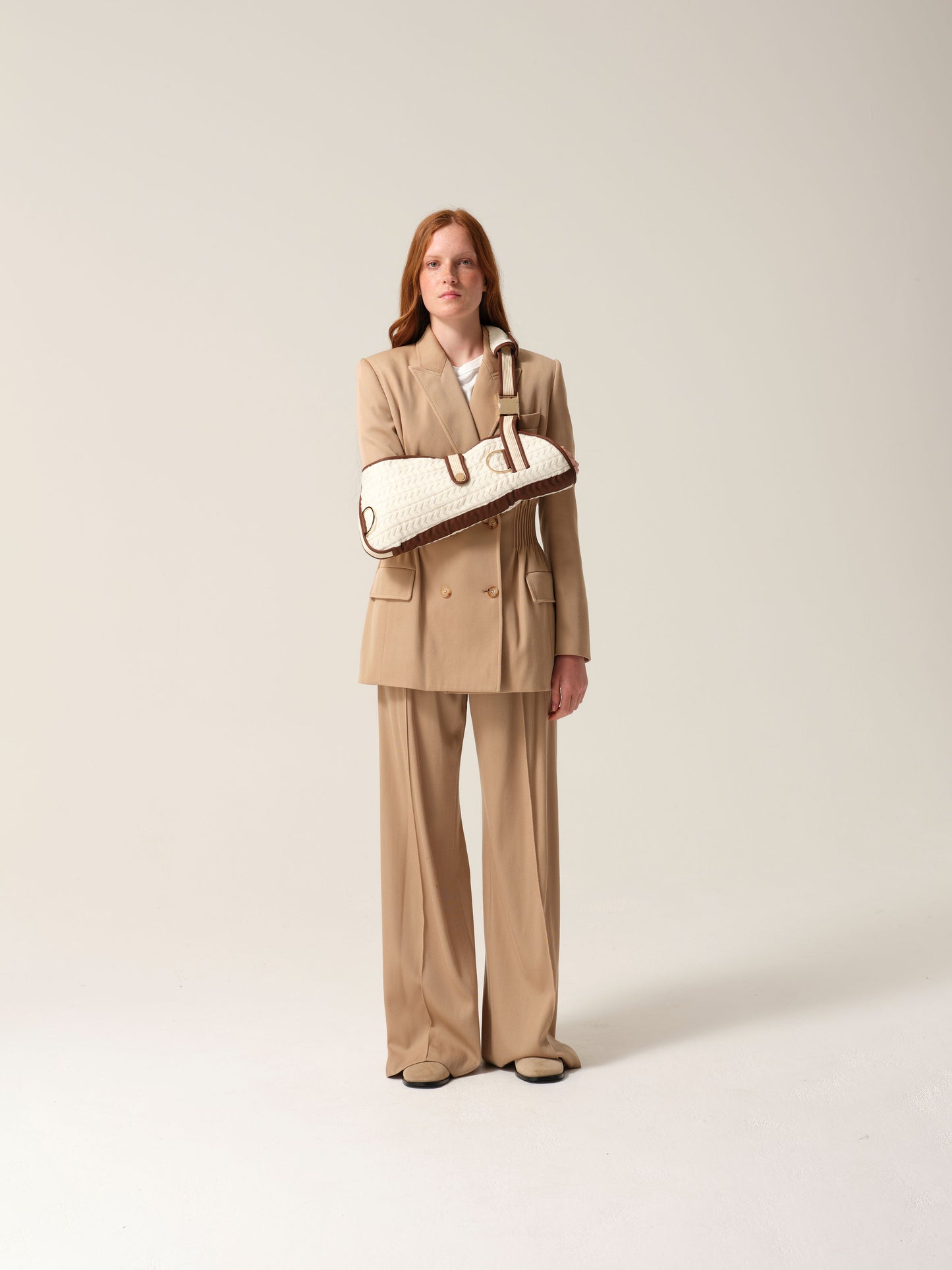 Woman wearing a camel suit with a white knitted cashmere arm sling shot against a plain background.