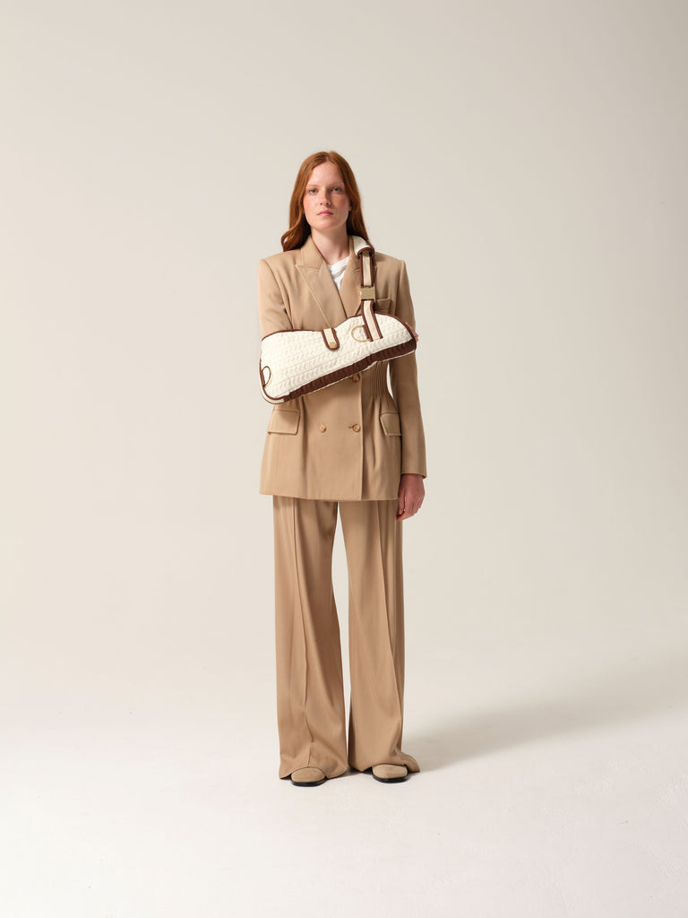 Woman wearing a camel suit with a white knitted cashmere arm sling shot against a plain background.