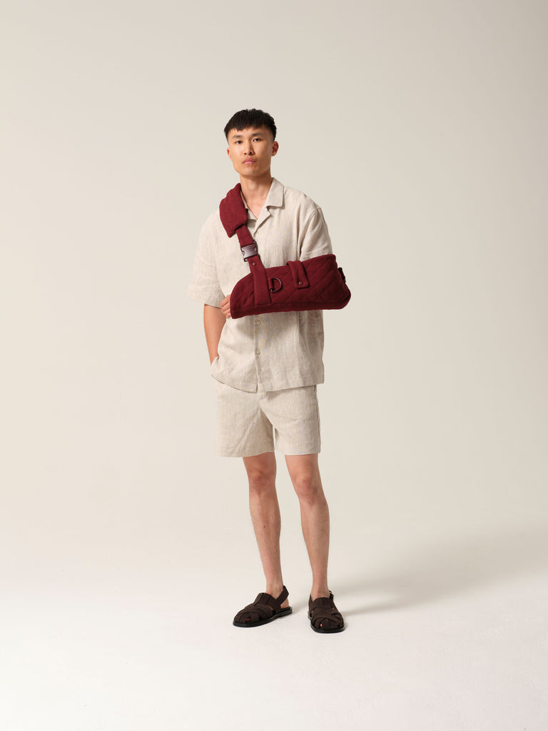 Man wearing a sand coloured linen shirtt and shorts with burgundy jersey arm sling featuring a wide shoulder strap and recycled plastic hardware against a plain background.