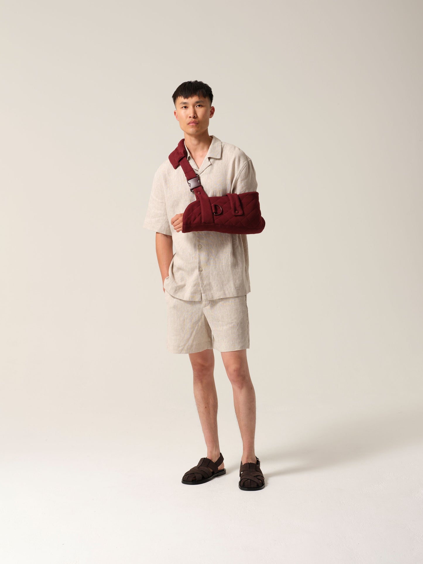Man wearing a sand coloured linen shirtt and shorts with burgundy jersey arm sling featuring a wide shoulder strap and recycled plastic hardware against a plain background.