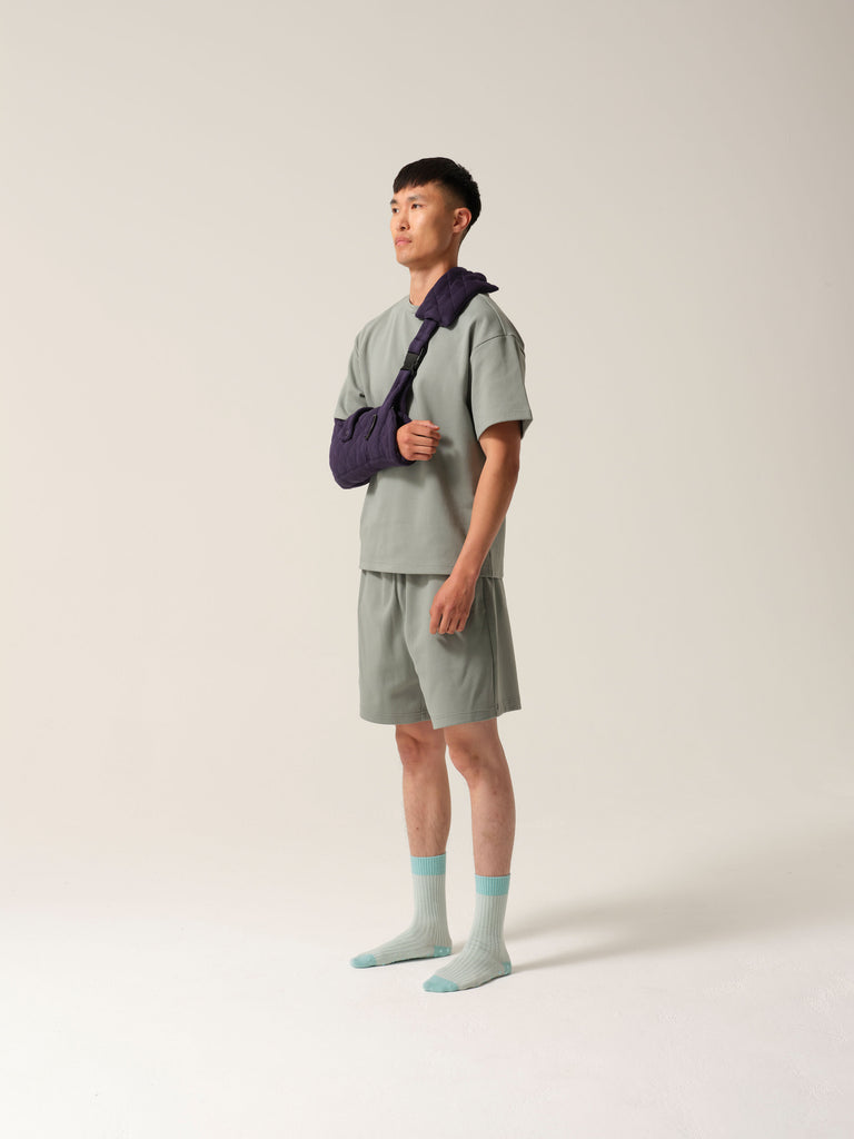 Man wearing a mint green tshirt and shorts weartng Navy jersey arm sling with a wide shoulder strap and recycled plastic hardware against a plain background with bofy tilted away from the camera.