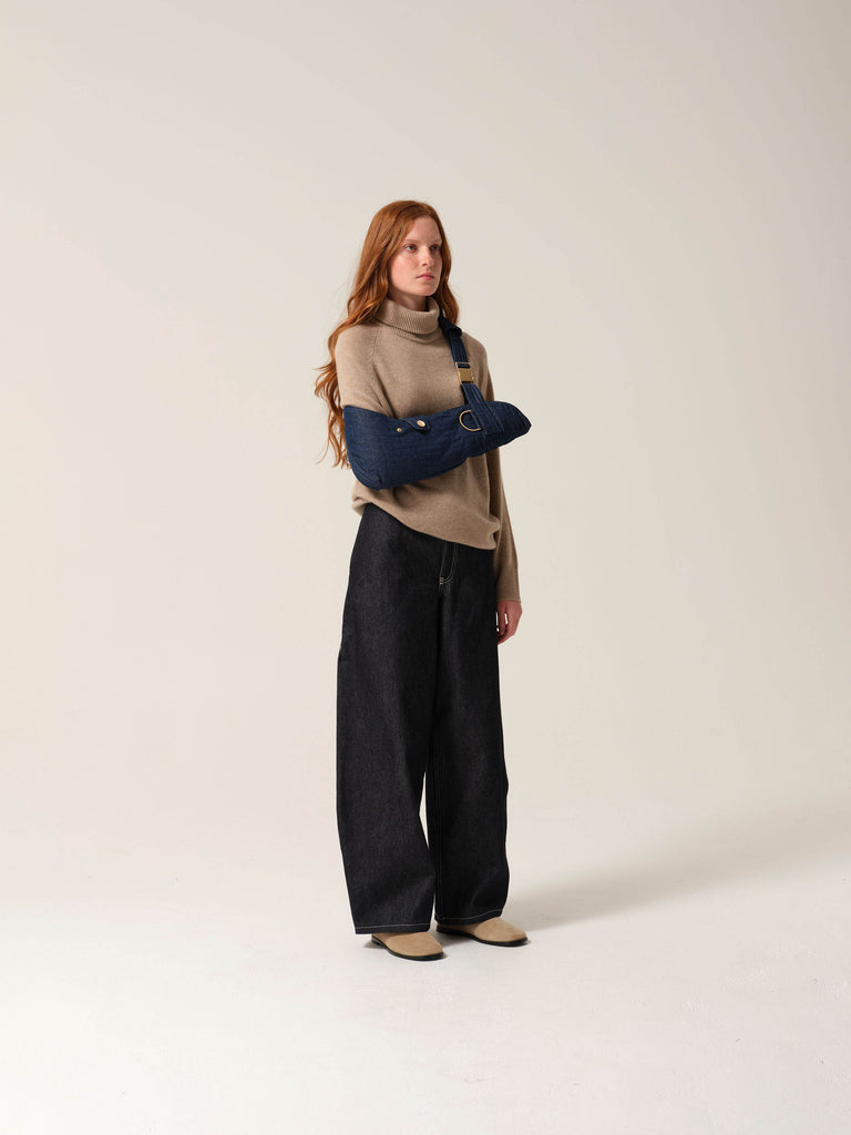 Woman wearing a beige sweater and black pants with a dark denim arm sling with gold metal work on one arm against a plain background.