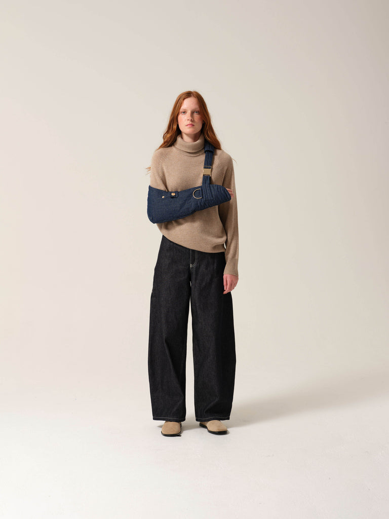 Woman wearing a beige sweater and black pants with a dark denim arm sling with gold metal work on one arm against a plain background.