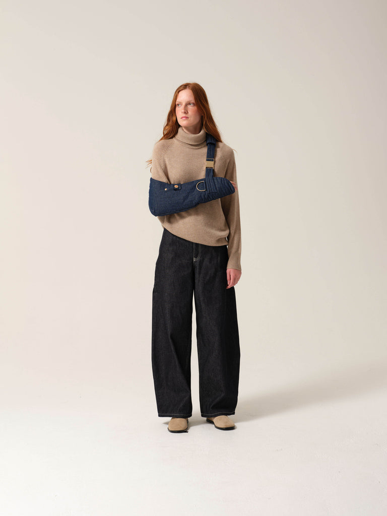 Woman wearing a beige sweater and black pants with a dark denim arm sling with gold metal work on one arm against a plain background.