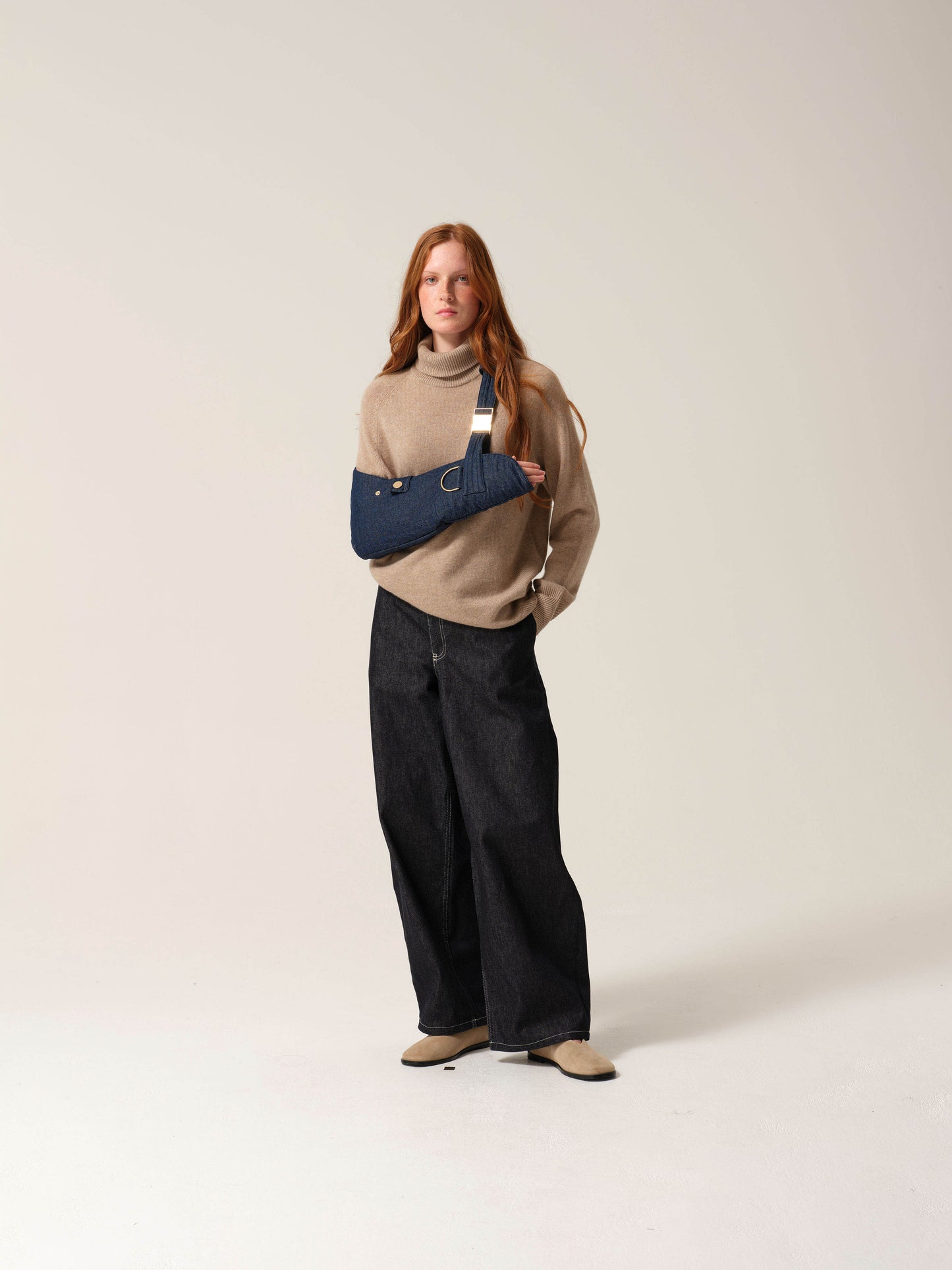 Woman wearing a beige sweater and black pants with a dark denim arm sling with gold metal work on one arm against a plain background.
