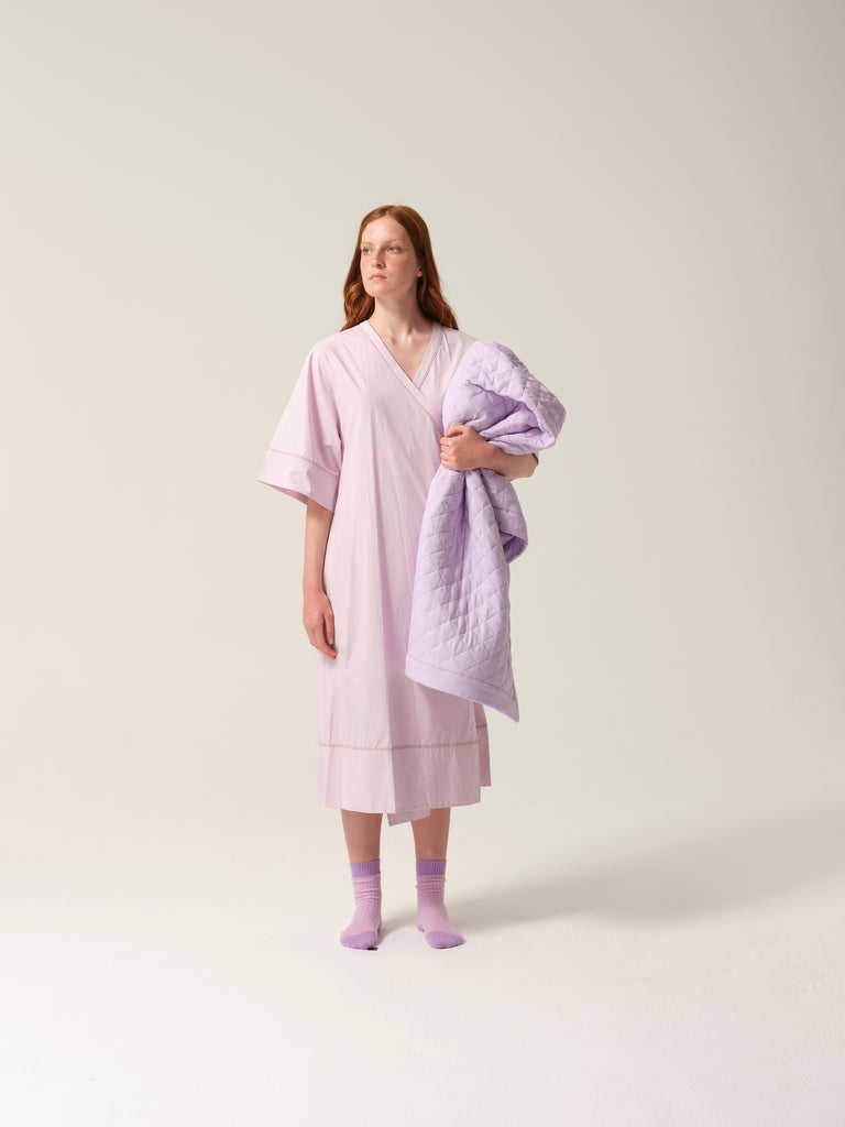 Woman wearing a light lilac patient robe holding a matching quilted blanket against a white background