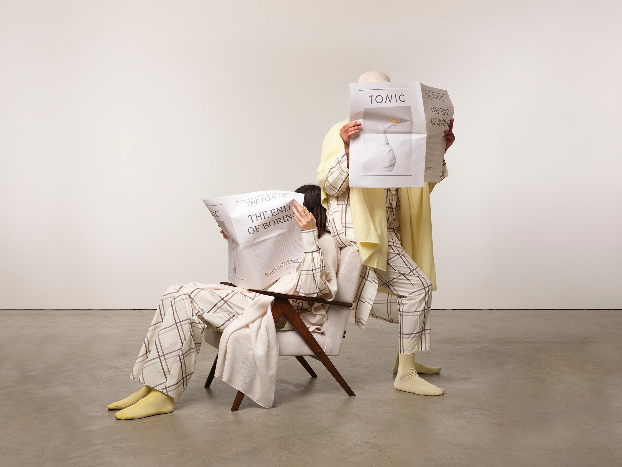 Image is of two models reading news papers one on chair and one standing while dressed in checked pyjamas with matching dressing gown and cashmere shawls. The colours are cream, silver grey and lemon yellow. This is recovery clothing for those recuperating from surgery or trreatment.