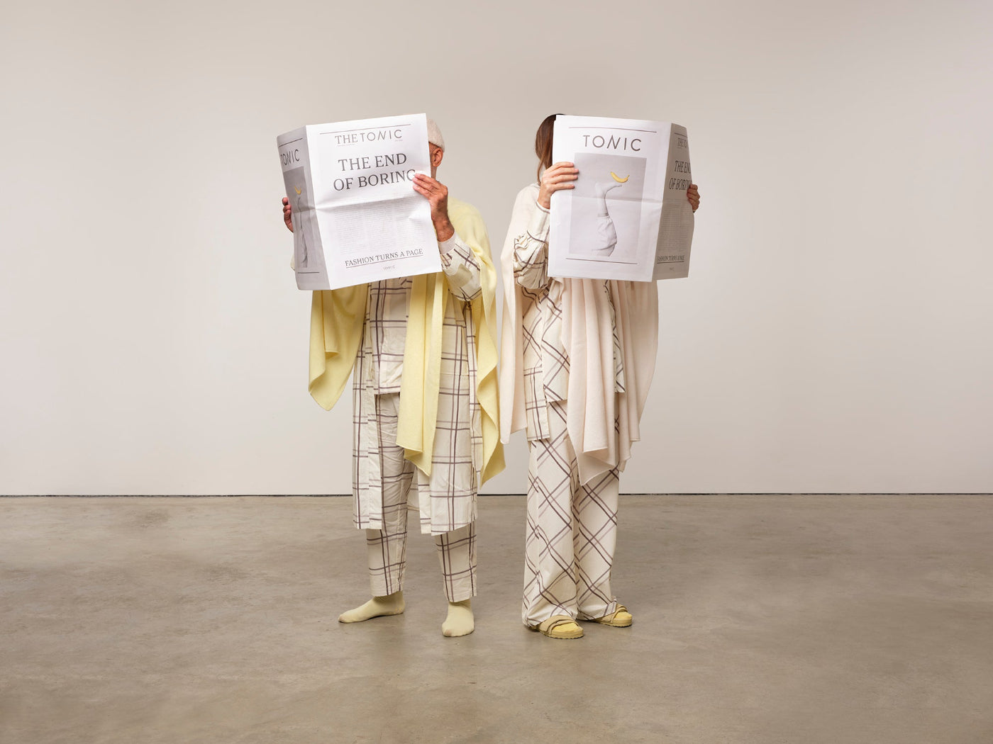 Image is of two models standing reading news papers while dressed in checked pyjamas with matching dressing gown and cashmere shawls. The colours are cream, silver grey and lemon yellow.