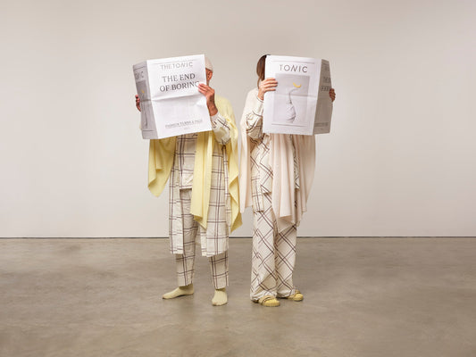 Image is of two models standing reading news papers while dressed in checked pyjamas with matching dressing gown and cashmere shawls. The colours are cream, silver grey and lemon yellow.