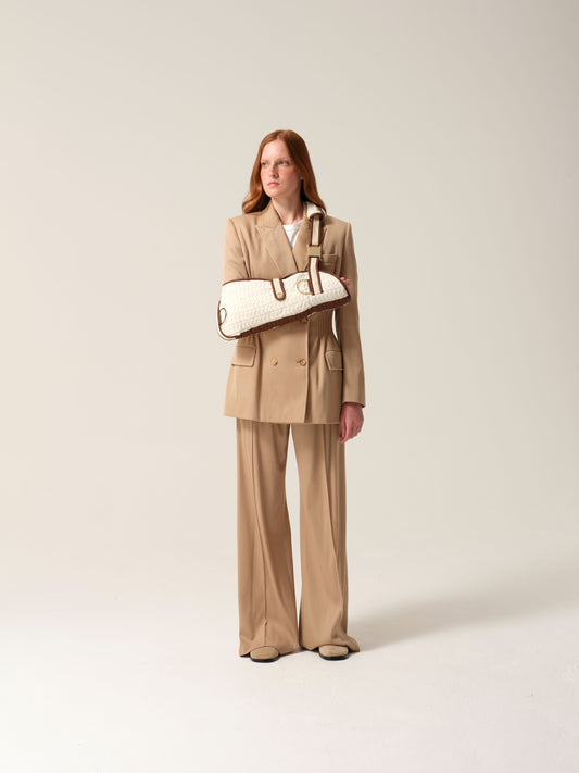 Woman wearing a camel suit with a white knitted cashmere arm sling shot against a plain background.