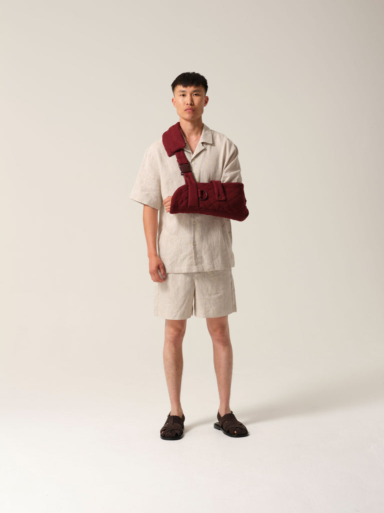 Man wearing a sand coloured linen shirtt and shorts with burgundy  jersey arm sling featuring a wide shoulder strap and  recycled plastic hardware  against a plain background.