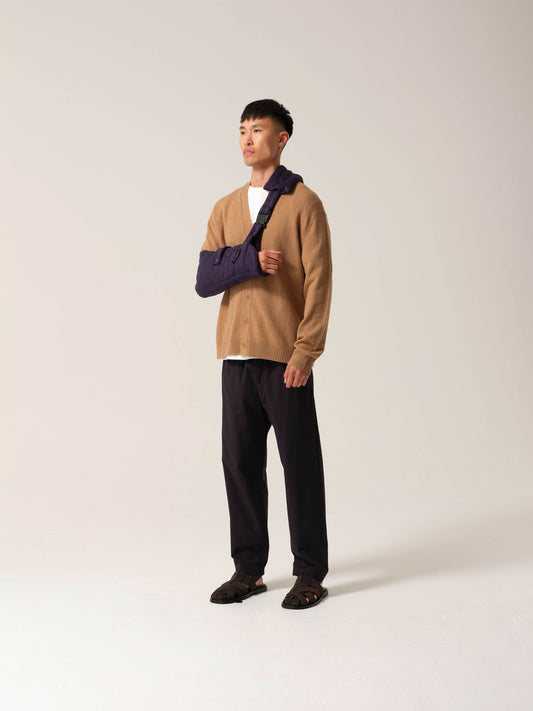 Person wearing a tan sweater and dark pants with a Navy jersey arm sling with a wide shoulder strap and recycled plastic hardware against a white background.