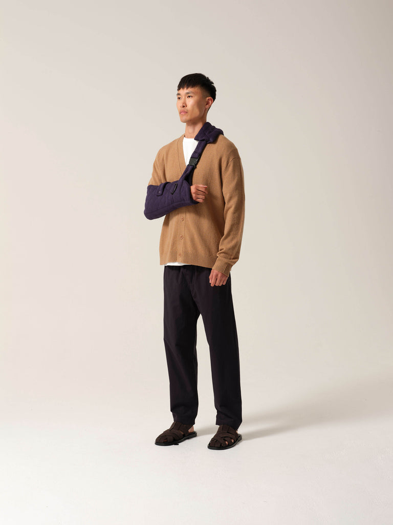 Person wearing a tan sweater and dark pants with a Navy jersey arm sling with a wide shoulder strap and recycled plastic hardware against a white background.