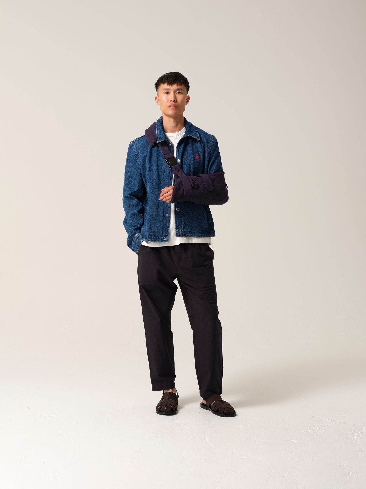 Man wearing a blue denim jacket with a white shirt and dark pants on a plain
background with navy jersey arm sling with a wide shoulder strap and recycled plastic hardware.
