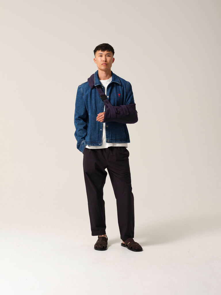 Man wearing a blue denim jacket with a white shirt and dark pants on a plain
background with navy jersey arm sling with a wide shoulder strap and recycled plastic hardware.