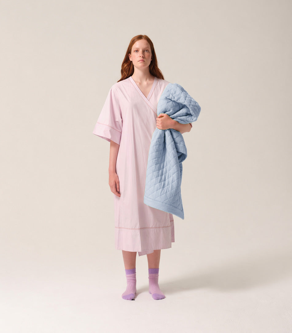 Woman in a pink patient gown holding a blue quilted blanket against a plain background