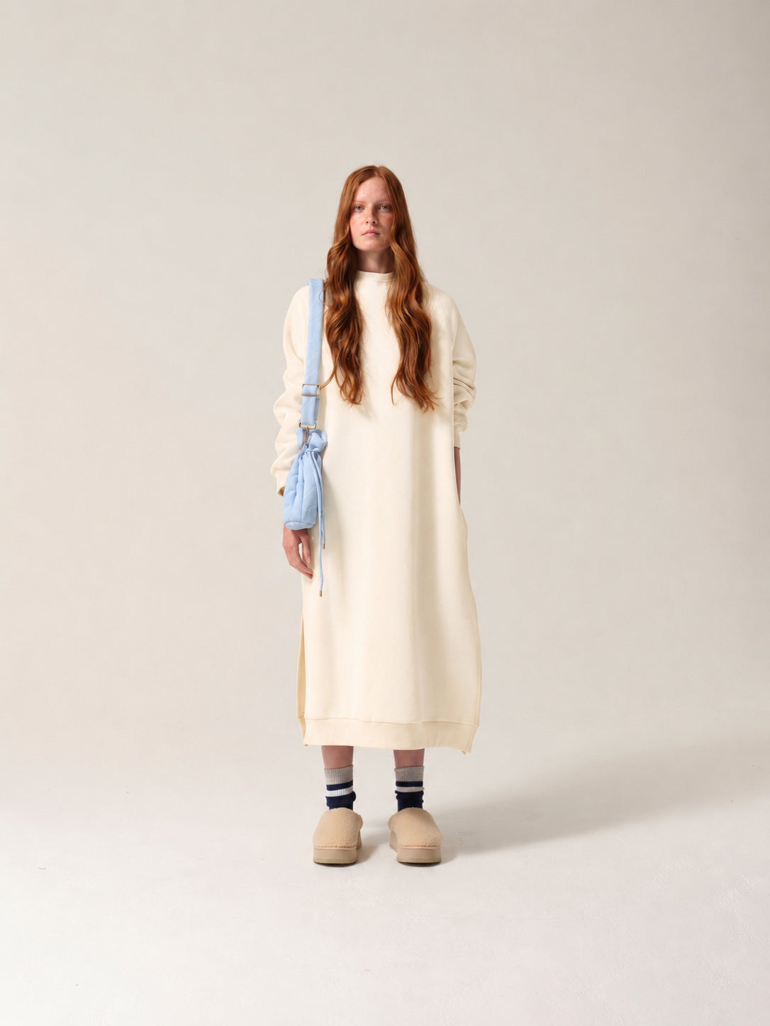 Woman wearing a long cream dress with a light blue drain bag against a plain background